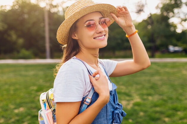 稻草戴着草帽 戴着粉色太阳镜 微笑着行走在公园里的年轻漂亮女人的肖像 夏日时尚的风格 五颜六色的时髦装扮眼镜微笑太阳镜