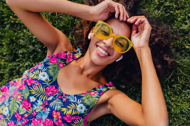 积极年轻时尚的黑人女子 戴着无线耳机听音乐 躺在公园的草地上玩得很开心 夏日的时尚风格 五颜六色的时髦装扮 俯瞰风景女孩太阳镜无线