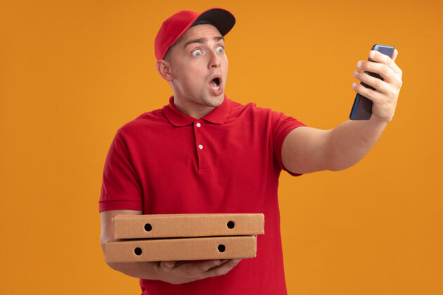 害怕害怕的年轻送货员穿着制服 戴着帽子 拿着披萨盒 看着他手里的电话 被隔离在橙色的墙上戴着帽子盒子