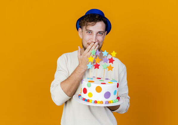 年轻年轻帅气的斯拉夫派对男 戴着派对帽 拿着生日蛋糕和明星们一起看着相机 手放在嘴巴上 在橙色背景上 留着复制空间派对橙色生日
