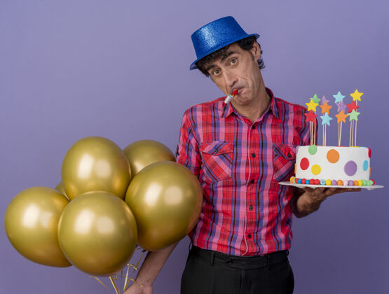 聚会令人印象深刻的中年白种人聚会男子戴着聚会帽拿着气球和生日蛋糕 嘴里叼着聚会吹风机 看着紫色背景上孤立的摄像机印象手捧吹风机