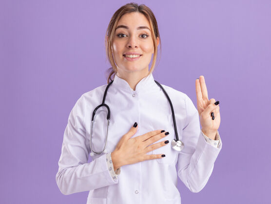 年轻令人高兴的年轻女医生穿着医用长袍 听诊器显示的姿势 隔离在紫色的墙上穿手势听诊器