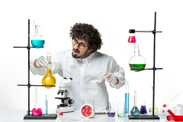 套装正面图身着特殊套装的年轻男性科学家站在浅白的墙上 拿着解决方案围着桌子实验容器烧杯