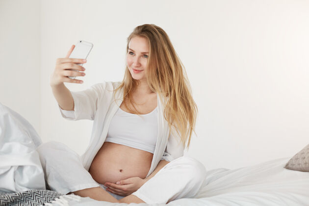 期待产前自拍怀孕的未来妈妈用智能手机微笑着抱着肚子自拍休闲休息房子
