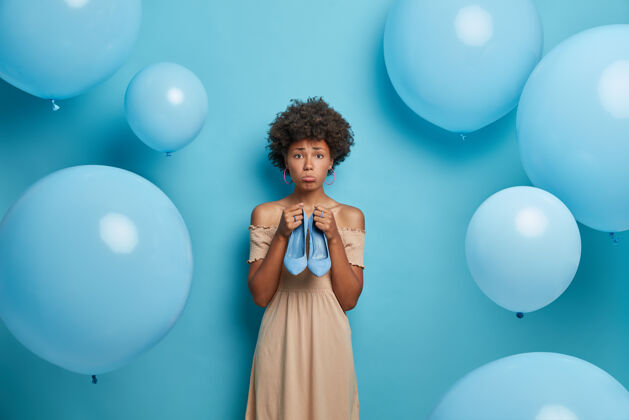 不高兴心烦意乱的黑皮肤卷发女子拿着蓝色高跟鞋 对着蓝色气球准备派对姿势姿势Buy美式