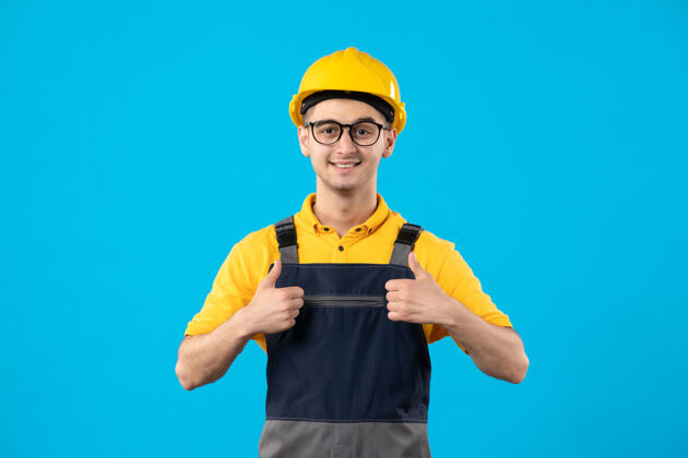 工人蓝色墙上穿着黄色制服的男建筑工人的正视图竖起大拇指视图专业