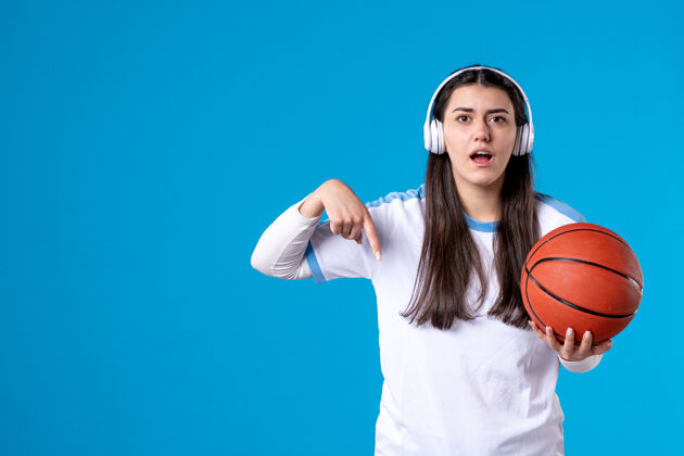 视图正面图：戴着耳机拿着篮球的年轻女性站在蓝色的墙上青年耳机手持