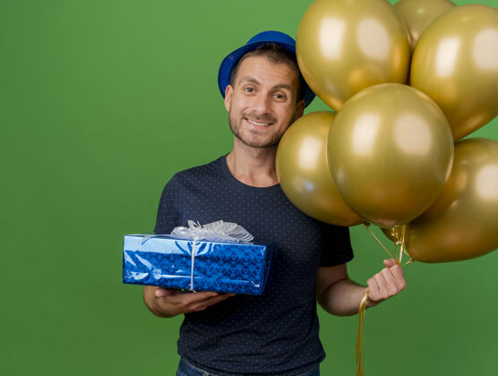 微笑微笑英俊的高加索男子戴着派对帽 手持氦气球和礼品盒 背景为绿色 并留有复印空间人礼物气球