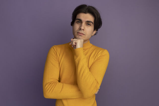 下巴自信的年轻帅哥穿着黄色高领毛衣把手放在下巴下面隔离在紫色的墙上穿高领毛衣毛衣