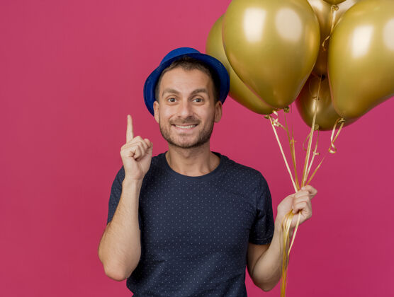 微笑面带微笑的白人帅哥戴着蓝色派对帽 手里拿着氦气球 在粉色背景上孤立地指向上 还有复制空间气球复制指向