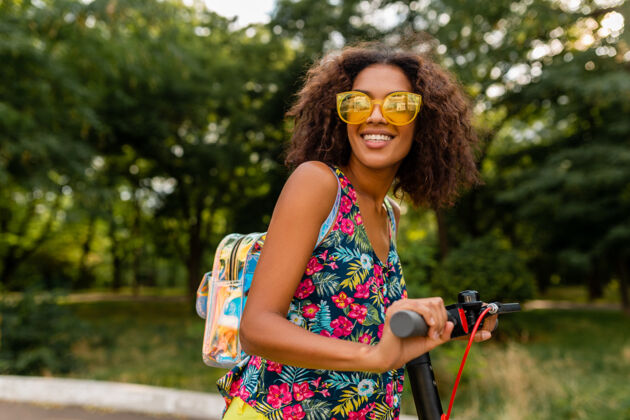 乘坐年轻时尚的黑人女子在公园里玩得很开心骑着电动脚踏车在夏天的时尚风格 五颜六色的时髦服装 穿着背包和黄色太阳镜女孩微笑时尚