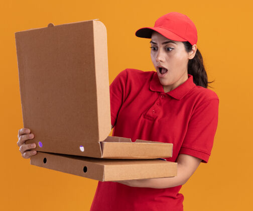 拿着惊讶的年轻送货女孩穿着制服 戴着帽子拿着披萨盒打开隔离在橙色墙上女孩送货盒子