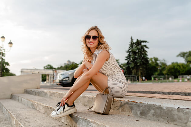 手提包年轻漂亮时尚的金发女郎坐在城市街道上 穿着夏日时尚的连衣裙 戴着太阳镜 钱包 银色运动鞋腿魅力裙子
