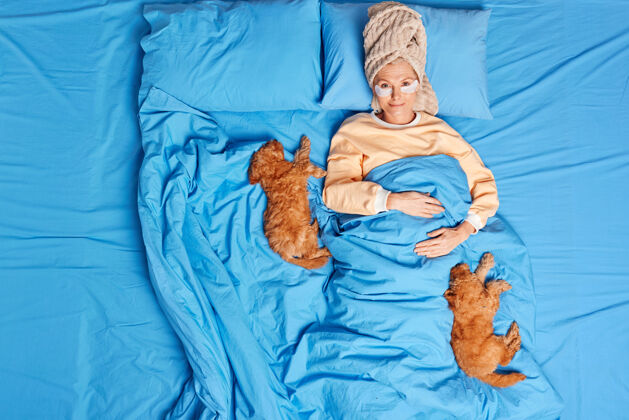 胶原蛋白上图：欧洲高级女士头戴睡衣裹毛巾眼罩减少皱纹与两只毛茸茸的小狗躺在床上 在舒适的卧室里接受美容治疗人们的生活方式美容枕头睡衣