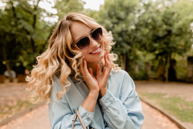 包迷人的金发美女穿着时髦的服装 戴着优雅的太阳镜和钱包在公园散步春天快乐微笑