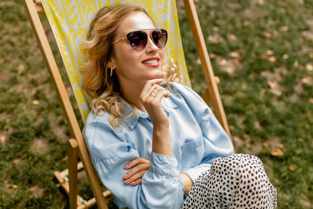 衣服迷人的金发微笑的女人 穿着时髦的服装 轻松地坐在躺椅上积极休闲包