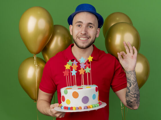 斯拉夫人面带微笑的年轻帅哥斯拉夫党人戴着党帽站在气球前拿着生日蛋糕看着相机做ok标志隔离在绿色背景上标志气球派对