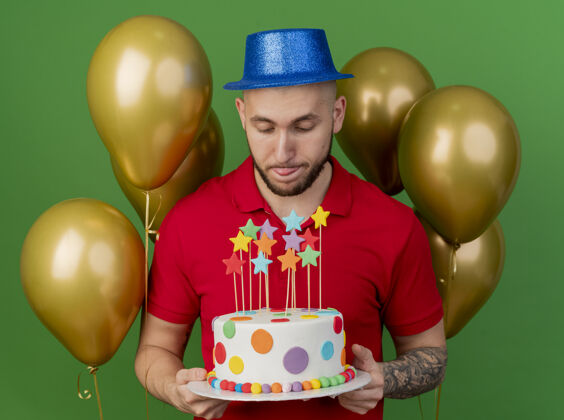 前面年轻英俊的斯拉夫人 戴着派对帽 站在气球前 拿着生日蛋糕 在绿色的背景上露出孤立的舌头生日蛋糕拿着