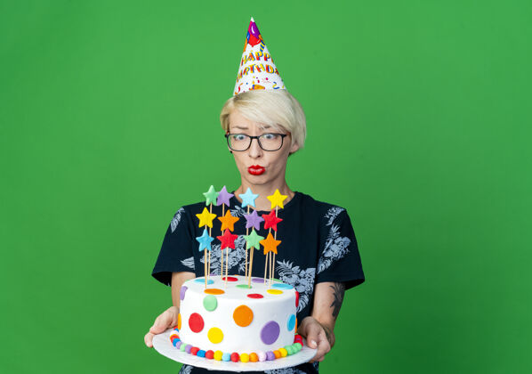 眼镜令人印象深刻的年轻金发派对女孩戴着眼镜和生日帽举行 看着生日蛋糕上的绿色背景与复制空间隔离派对空间印象