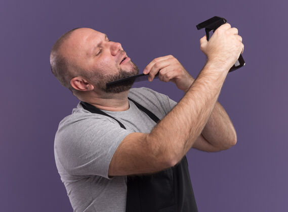 梳子高兴的斯拉夫中年男理发师穿着制服梳理胡须 用紫色墙上隔离的喷雾瓶浇水胡须男请