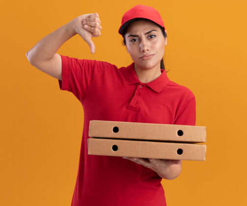 穿穿着制服 戴着帽子 拿着披萨盒 拇指朝下的年轻送货女孩被隔离在橙色的墙上放下盒子年轻