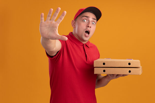 显示被吓坏了的年轻送货员穿着制服 戴着帽子 拿着披萨盒 在橙色的墙上显示着停止的手势停止盒子送货