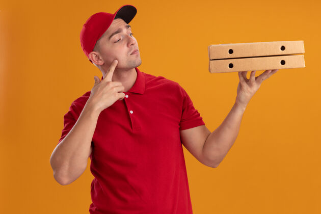 送货令人印象深刻的年轻送货员穿着制服 戴着帽子 看着披萨盒把手指放在脸颊上隔离在橙色的墙上拿着手指男人