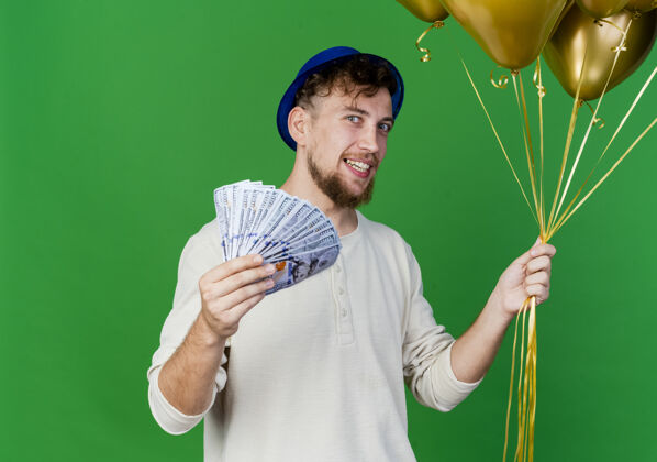 气球面带微笑的年轻英俊的斯拉夫党人戴着党的帽子拿着气球和钱看着相机隔离在绿色背景与复制空间钱斯拉夫人年轻人