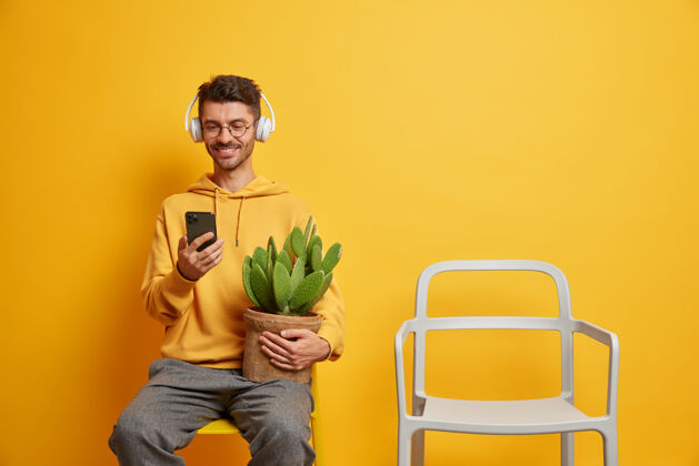 男人快乐的家伙用手机在播放列表中下载音乐在耳机中听喜爱的歌曲在家里享受空闲时间在空椅子旁摆姿势设备人放松