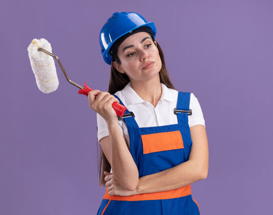 女人印象深刻的看着身边穿着制服的年轻建设者女子拿着滚筒刷隔离在紫色的墙上年轻人滚轴壁板
