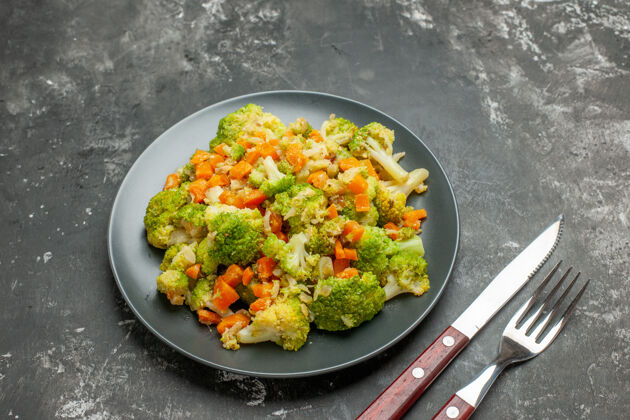 午餐健康餐 黑盘子上放花椰菜和胡萝卜 灰色桌子上放刀叉锅厨具健康餐