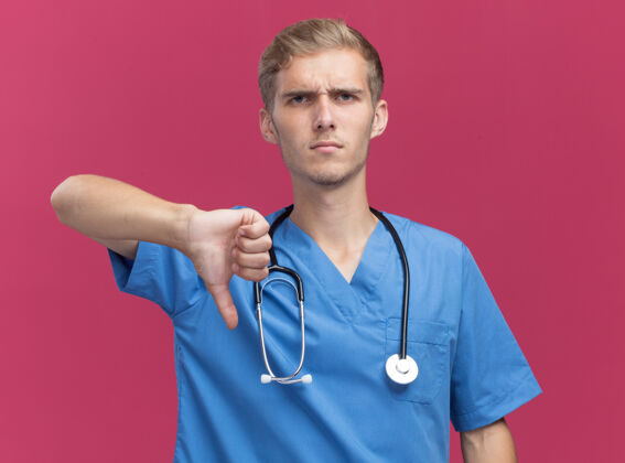 穿年轻的男医生穿着医生制服 听诊器显示拇指朝下 隔离在粉红色的墙上不愉快制服向下