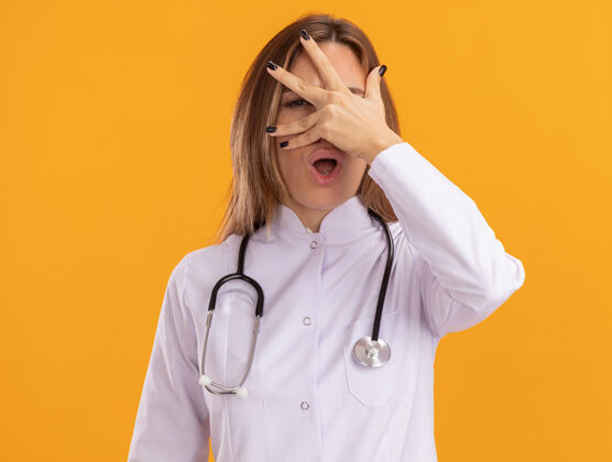 惊喜惊讶的年轻女医生穿着医用长袍 听诊器捂着脸 手被隔离在黄色的墙上穿医生女性
