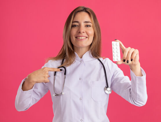 女性面带微笑的年轻女医生穿着医用长袍 手持听诊器 指着隔离在粉红色墙上的药片抱听诊器年轻