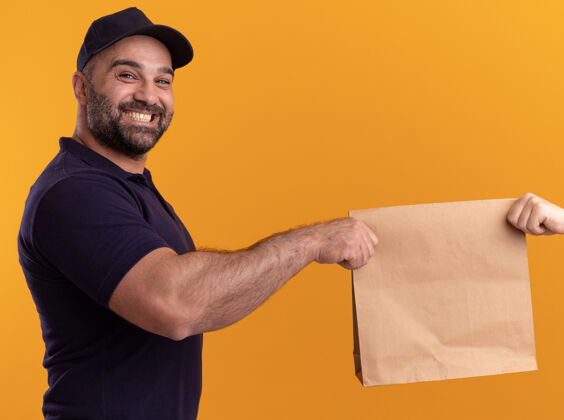 顾客微笑的中年送货员 穿着制服 戴着帽子 把纸食品包装交给隔离在黄色墙上的客户纸张中年微笑
