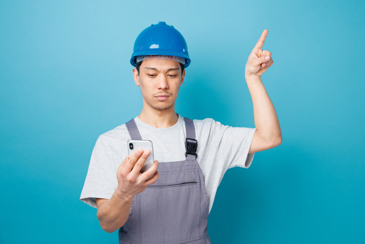 蓝色严肃的年轻建筑工人戴着安全帽 穿着制服 手持并看着手机指着上面头盔电话拿着