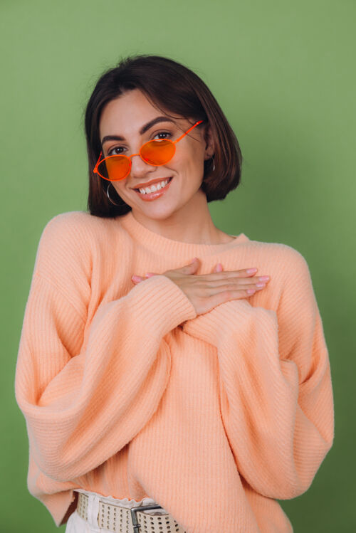 爱年轻时尚的女士穿着休闲桃色毛衣 戴着橘色眼镜 隔着绿橄榄色的墙正面双手合十放在胸前 心复制空间面部时尚温柔