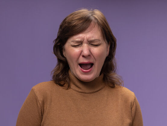 女人沮丧的中年妇女穿着棕色高领毛衣 闭着眼睛站在紫色的墙上痛哭站闭嘴高领毛衣