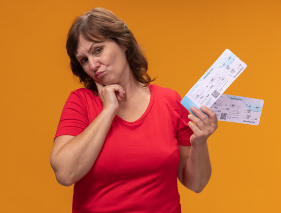 中年身穿红色t恤的中年妇女站在橙色的墙上 手里拿着机票 表情怀疑怀疑立场飞机