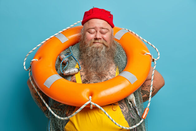 帽子快乐快乐的大胡子渔夫站着闭着眼睛提着橙色充气的救生圈在渔船上悠闲地摆姿势男人粗壮救生圈