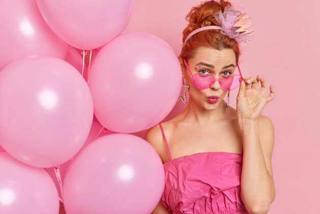 充气美丽迷人的红发魅力的特写镜头年轻女子看起来从时髦的粉红色太阳镜下穿着时髦的服装举行氦气球站在室内庆祝生日氦皇冠头发