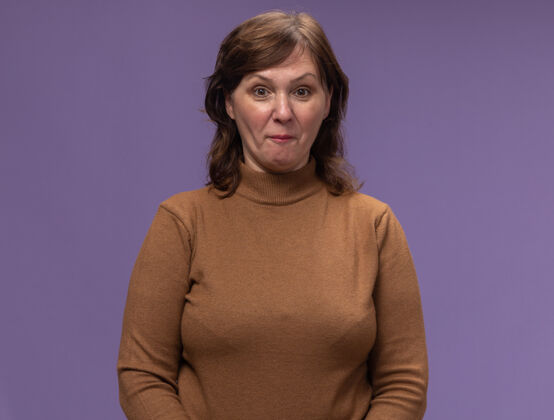 中年穿着棕色高领毛衣的中年妇女站在紫色的墙上感到困惑和惊讶女人惊讶站