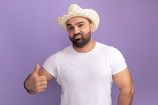 表情胡子男人穿着白色t恤 戴着夏日帽 自信地站在紫色的墙上竖起大拇指男人拇指胡子