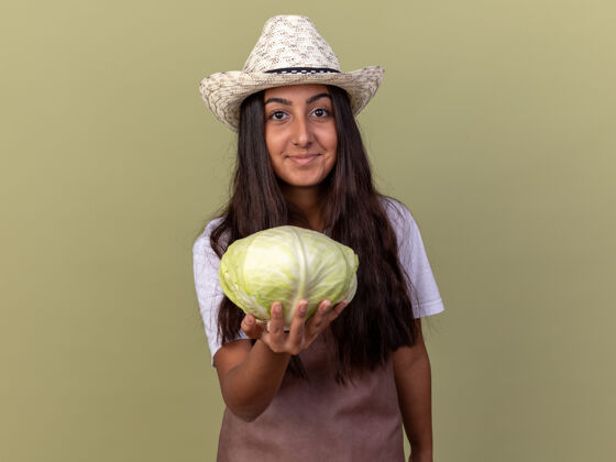 年轻穿着围裙 戴着夏帽 脸上挂着微笑的白菜的年轻园丁女孩站在绿色的墙上站起来围裙花园