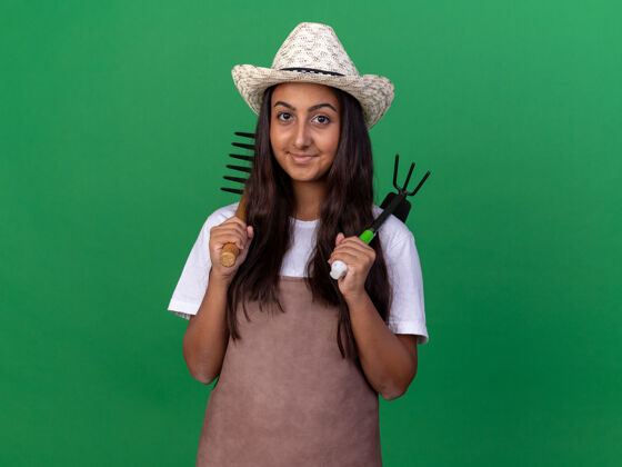 帽子快乐的年轻园丁女孩 围裙 戴着夏帽 手里拿着小耙子 面带微笑地站在绿色的墙上围裙微笑年轻