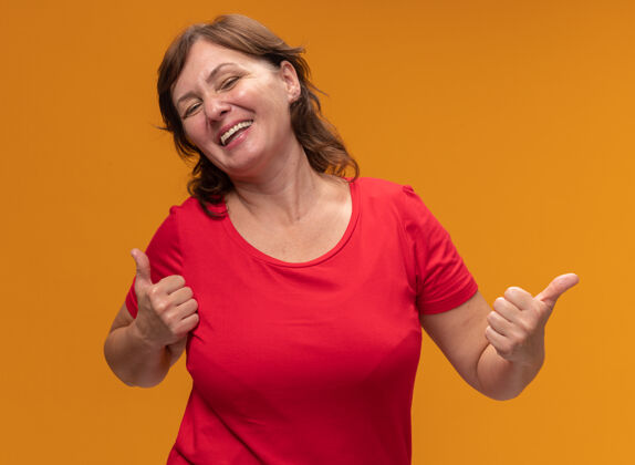拇指身穿红色t恤的中年妇女站在橘色的墙上 高兴地竖起大拇指女人中间表演