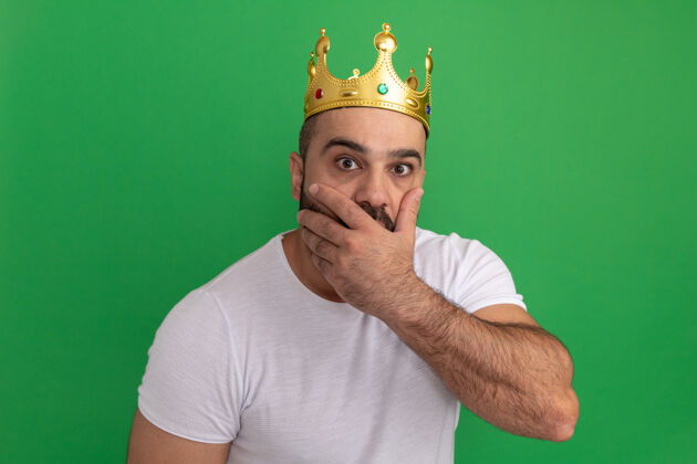 男人戴着金色王冠的白t恤胡子男人被震惊了 用手捂住嘴站在绿色的墙上掩护胡子皇冠