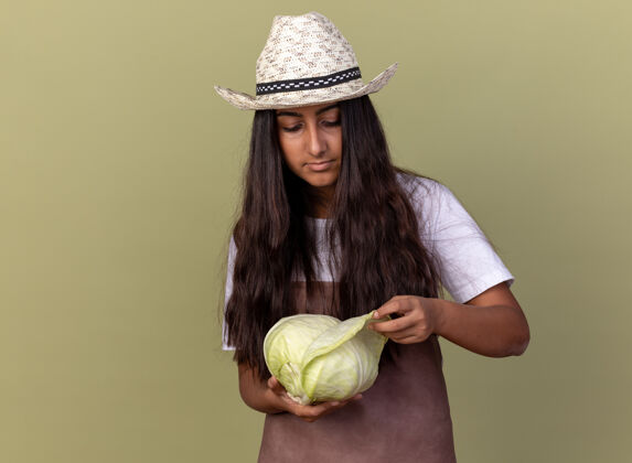 立场年轻的园丁女孩围着围裙 戴着夏帽 手里拿着卷心菜 站在绿色的墙边好奇地看着它花园卷心菜围裙