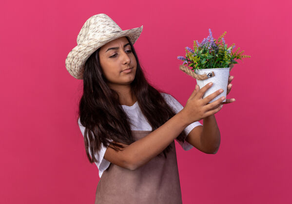 花盆年轻的园丁女孩围着围裙 戴着夏帽 手里拿着盆栽植物 站在粉色的墙上好奇地看着它看台帽子年轻人