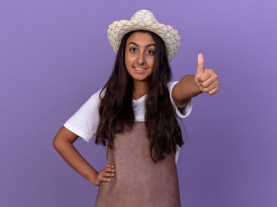 年轻人穿着围裙 戴着夏帽的年轻园丁女孩站在紫色的墙上 愉快地微笑着竖起大拇指花园欢呼看台